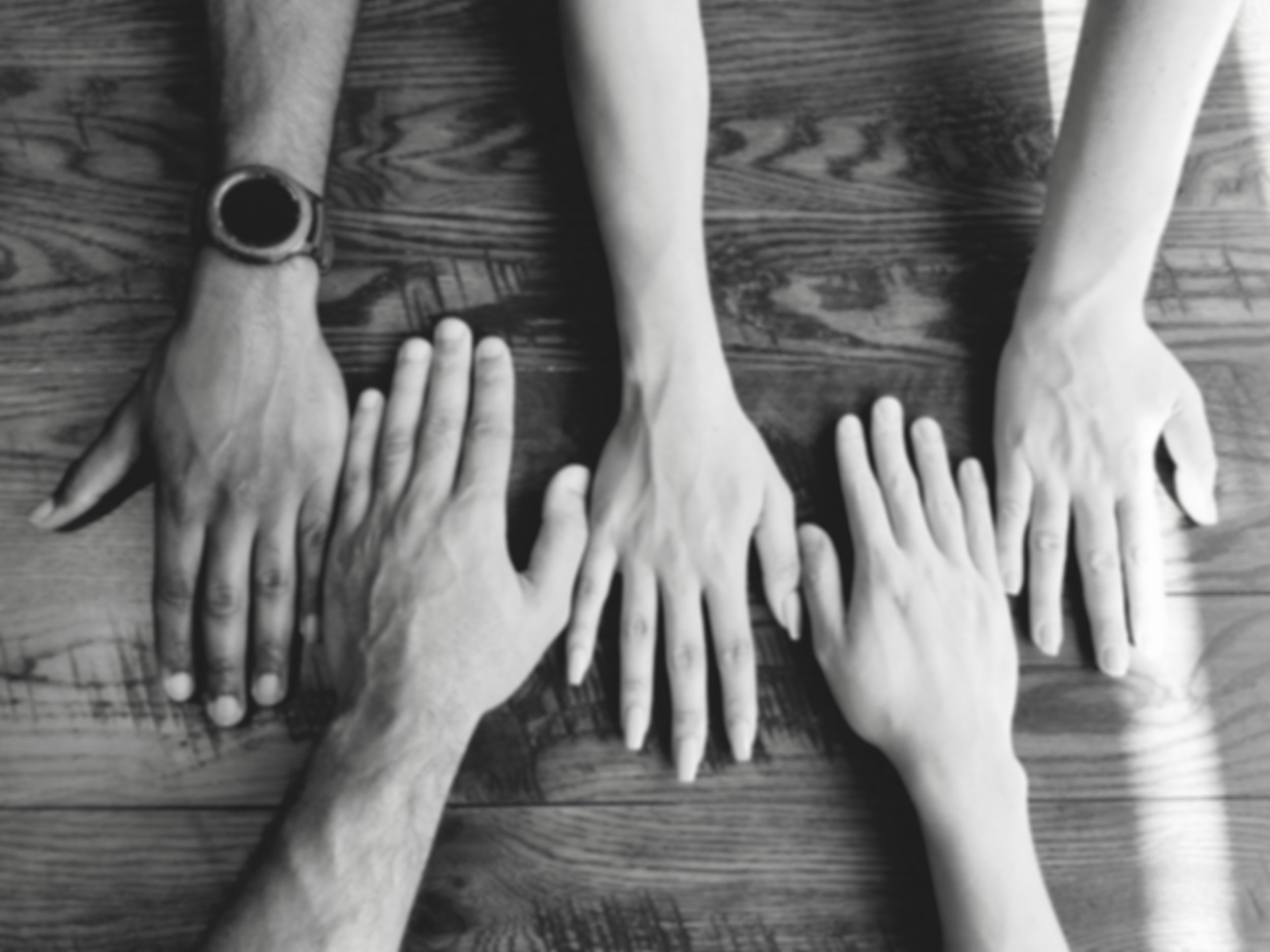 Five people's hands reaching together toward the center of a wooden table, symbolizing partnership